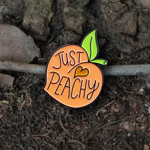 Détail d'un pin's pêche en émail avec texte Just Peachy sur un fond en bois naturel