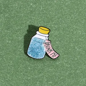 Detail view of a glittery blue potion bottle enamel pin with a Drink Me tag on green paper