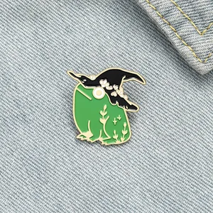 Close-up of a cute green frog enamel pin wearing a black witch hat on denim fabric