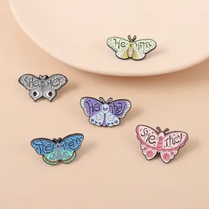 Five butterfly pronoun enamel pins in various colors on a soft cream background