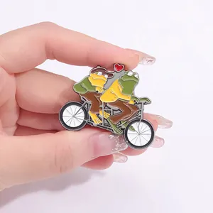 Détail d'un pin's en émail représentant des grenouilles à vélo tenu à la main sur fond rose