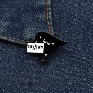 Close-up of crow enamel pin with THEY/THEM text on a blue denim jacket background