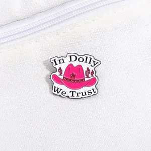 Close-up of pink enamel cowboy hat pin with 'In Dolly We Trust' text on canvas