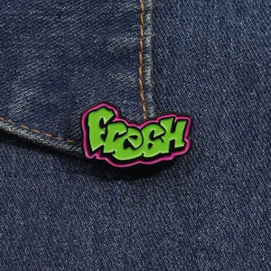 Nahaufnahme der grünen Graffiti-Emaillenadel 'Fresh' auf dunkelblauem Jeansstoff