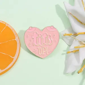 Detail of pink heart enamel pin with gold trim and tits UP lettering on a green surface