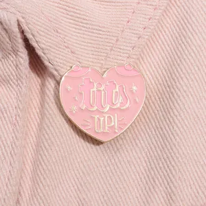 Close-up of a pink heart-shaped enamel pin with tits UP! text on pink denim fabric