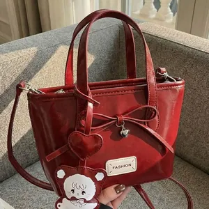 Dunkelrote Lackleder-Eimertasche mit Schleife und Bärenanhänger auf grauem Sofa