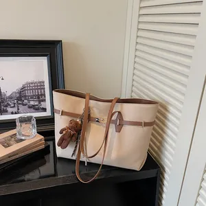Large beige tote bag with brown trim and cute plush bear charm, minimalist indoor scene