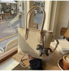 Detalle de un bolso tipo cubo de lona beige con ribete de cuero y dije de caballo