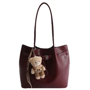 Burgundy faux leather tote bag with long straps and a plush bear charm on white background