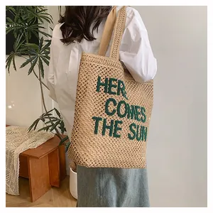 Detail of beige woven tote bag with green 'HERE COMES THE SUN' knit text and wide strap