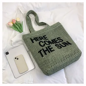 Close-up of green woven beach tote featuring HERE COMES THE SUN slogan on white fabric