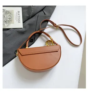 Close-up of brown leather saddle bag featuring unique large gold ring handle on a white surface