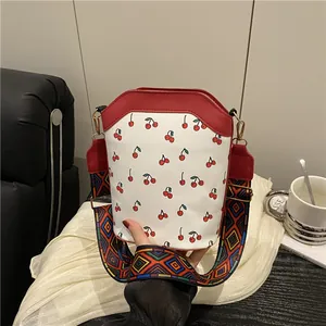 White cherry print bucket bag with red trim and wide geometric woven shoulder strap