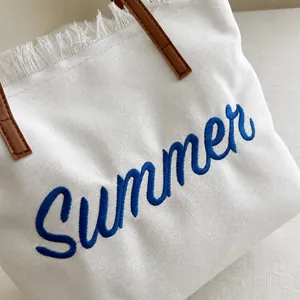 Detail of white canvas tote featuring blue towel embroidery and brown leather handles