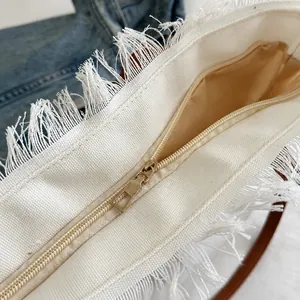 Close-up of white canvas tote bag featuring fringe trim and premium gold zipper detail
