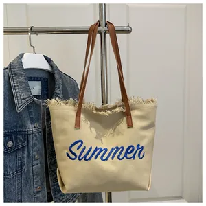 Detalle de bolso tote de lona beige con bordado Summer azul y bordes de flecos finos