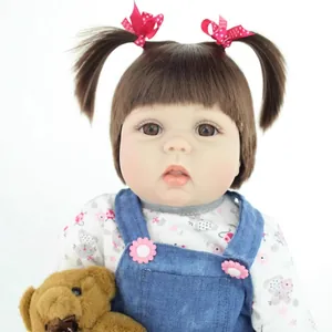 Lifelike reborn baby doll with brown pigtails holding a teddy bear in denim overalls