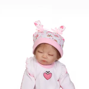 Lifelike reborn baby doll with floral hat and pink heart top on white background