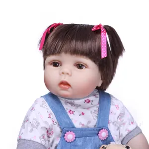 Lifelike baby girl doll with brown hair and denim pinafore dress on white background