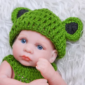 Lifelike 26cm reborn baby doll wearing a green frog knitted hat on white plush background