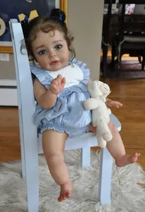 Close-up of 60cm reborn baby doll with blue eyes, dark curls and plush bear on chair
