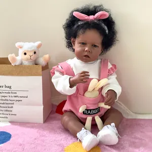 Muñeco Reborn Liam de piel oscura con pelo rizado, peto rosa y zapatos blancos de bebé