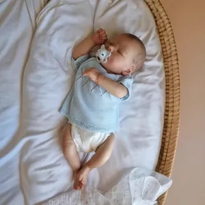 Lifelike 16-inch reborn baby doll in blue knit shirt lying in a woven basket with pacifier