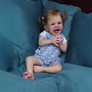 Poupée bébé réaliste de 60 cm en salopette fleurie assise sur un canapé en velours