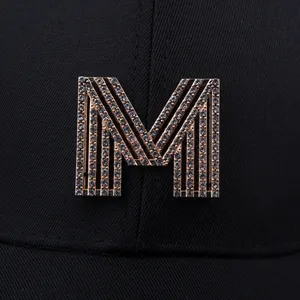 Detail eines strassbesetzten roségoldenen M-Emblems auf schwarzer Twill-Baseballkappe