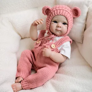 Handmade 19-inch reborn doll wearing pink knit overalls and a bear ear hat on plush background