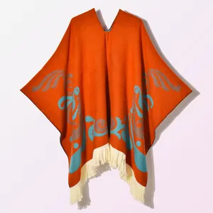 Detail of orange knitted poncho featuring intricate ethnic patterns and creamy white tassels