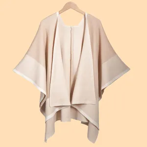 Elegant beige knitted shawl poncho with white contrast piping on a minimalist wood hanger