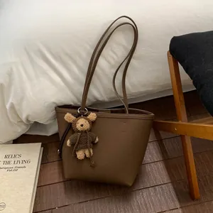 Detail of a brown leather tote bag featuring a cute plush bear pendant and thin shoulder straps