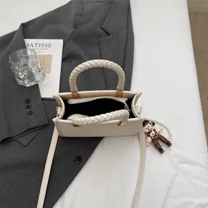 Elegante borsa quadrata crema con manico intrecciato e ciondolo su sfondo di classe