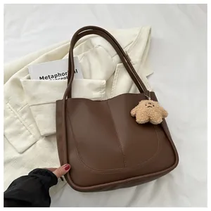 Detail of a brown PU leather tote bag featuring U-shaped stitching and a plush bear charm