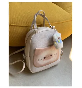 Detail of a beige cat-shaped backpack with fabric pocket and plush charm on a sofa