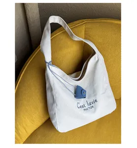 Detail of white canvas tote bag featuring blue C'est lavie embroidery and leather charm