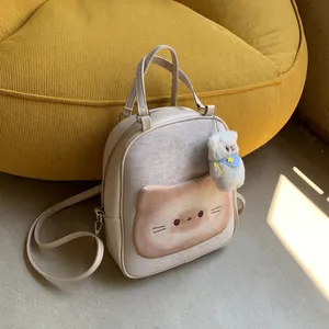 Cute beige cat-themed canvas backpack with cat embroidery and plush pendant on yellow sofa