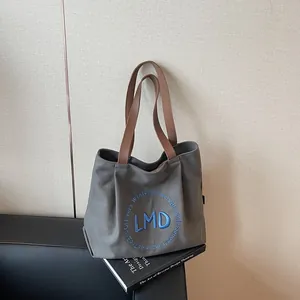 Grey canvas tote bag with brown straps and blue embroidery on a black leather sofa