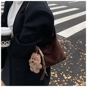 Brown suede shoulder bag with plush bear charm worn by a model on a street background