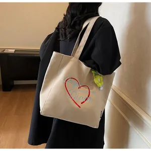 Detail der beigen Segeltuch-Tasche mit Herzstickerei und grünem Anhänger am Modell