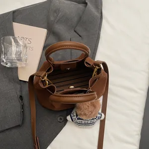 Brown pleated tote bag with bear charm and adjustable strap on a grey blazer backdrop