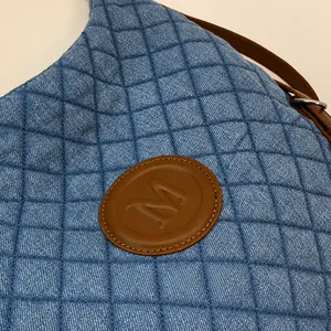 Close-up of quilted blue denim tote bag with leather M logo and brown shoulder strap
