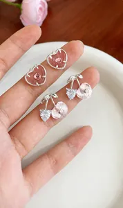Détail de boucles d'oreilles cœur et cerise rose sur le doigt montrant la petite taille