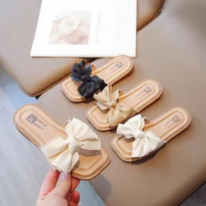 Girls' soft-sole bow slides in black and beige displayed on a leather stool