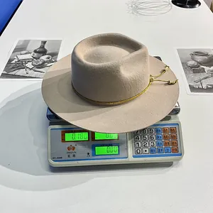 Dettaglio di cappello fedora in lana beige con cordino dorato su bilancia che segna 180g