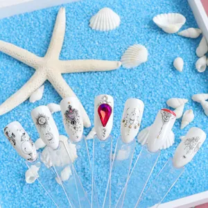 Dettaglio di unghie bianche con gemme viola e perle su sfondo con sabbia e conchiglie