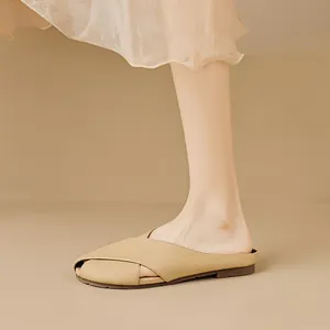 Κομψά μπεζ flat mules με χιαστί λουράκια φορεμένα με φούστα σε μίνιμαλ σκηνικό