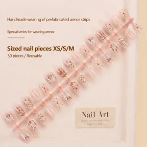 Handmade nude press-on nails with 3D gold bows, pearls, and glitter on beige leather